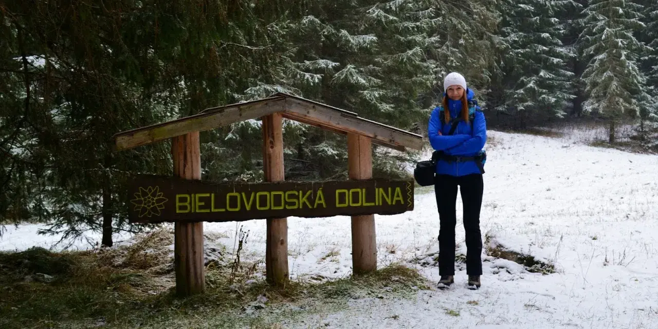 Kobieta w niebieskiej kurtce stoi przy drewnianym znaku "Bielovodská dolina" w zimowej scenerii.
