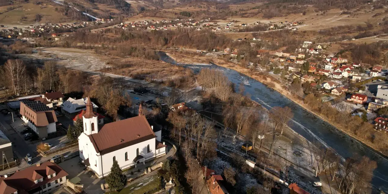 Widok z lotu ptaka na malowniczą dolinę Popradu zimą. Biały kościół z czerwoną wieżą góruje nad zabudowaniami.