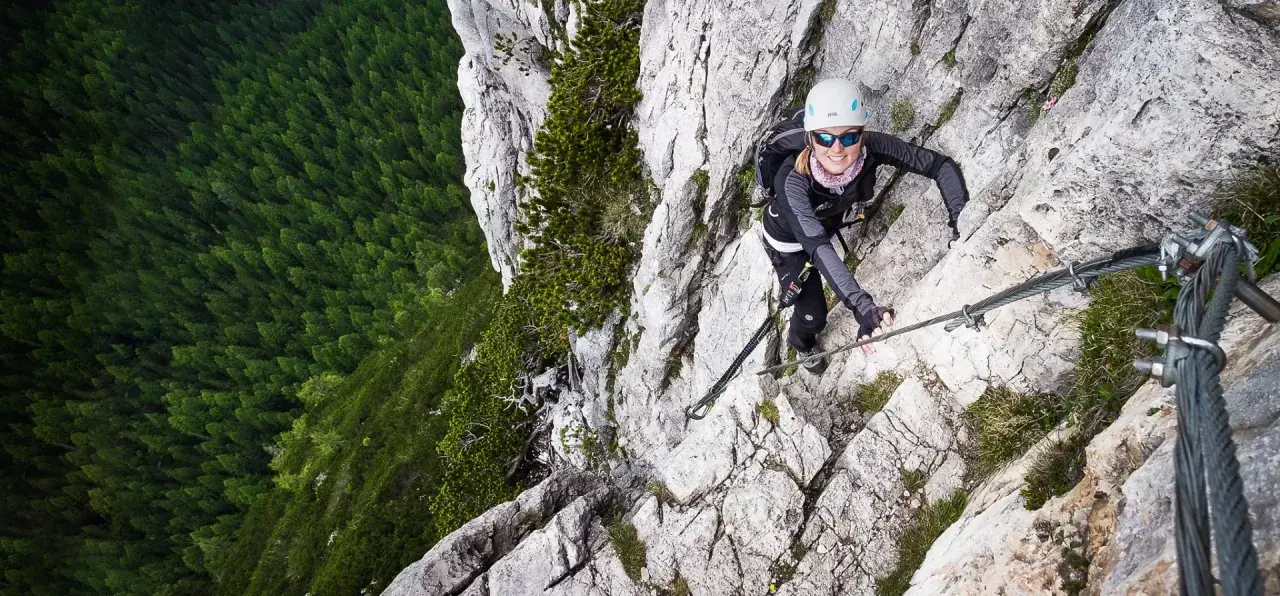 Kobieta wspina się po via ferrata w Dolomitach, z widokiem na gęsty las poniżej.