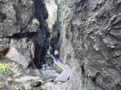 Wąska ścieżka przez skalny kanion w Małej Fatrze. Turyści idą drewnianym mostkiem wzdłuż strumienia.