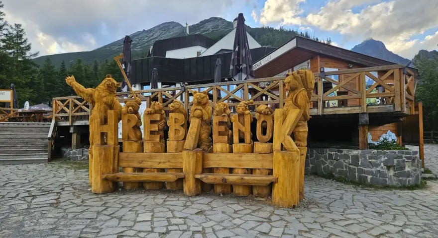 Drewniane rzeźby niedźwiedzi tworzą napis HREBENOK. W tle budynek z tarasem i góry. Sprawdź hrebienok kolejka cennik.