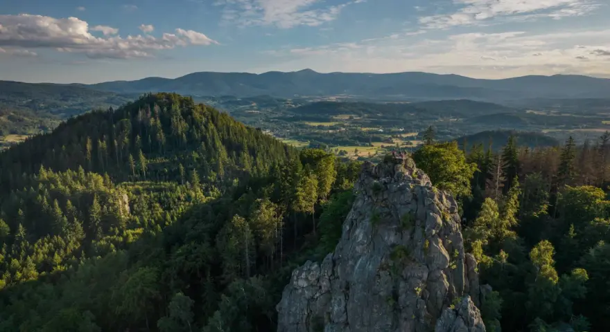 Widok na skalne formacje Rudawy Janowickie, porośnięte lasem, z ludźmi na szczycie. W tle rozległa panorama gór i dolin.