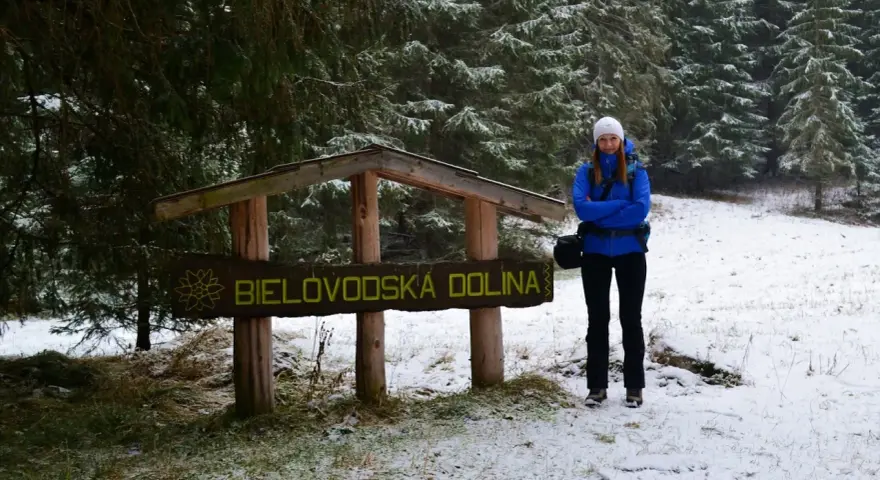 Kobieta w niebieskiej kurtce stoi przy drewnianym znaku "Bielovodská dolina" w zimowej scenerii.