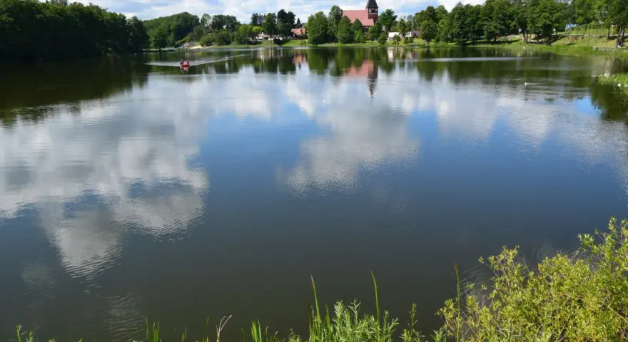 Spokojne jezioro na Kaszubach, odbijające chmury. W oddali widać łódkę i kościół. Idealne miejsce na wakacje.