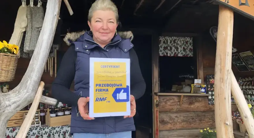 Kobieta z certyfikatem "Przejściowa Firma" przed bacówką. W tle widać rękodzieło i przetwory, a także tablicę z napisem "Bacówka u Jancora".