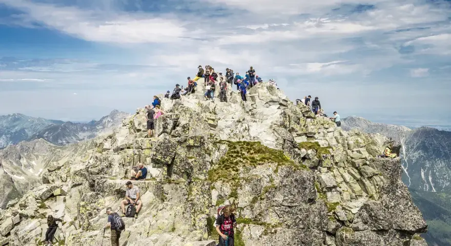 Tłum turystów na skalistym szczycie Rysów, z widokiem na Giewont w oddali.