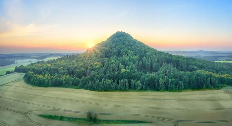 Wygasłe wulkany Dolny Śląsk o poranku. Słońce wschodzi nad zalesionym stożkiem, otoczonym polami.