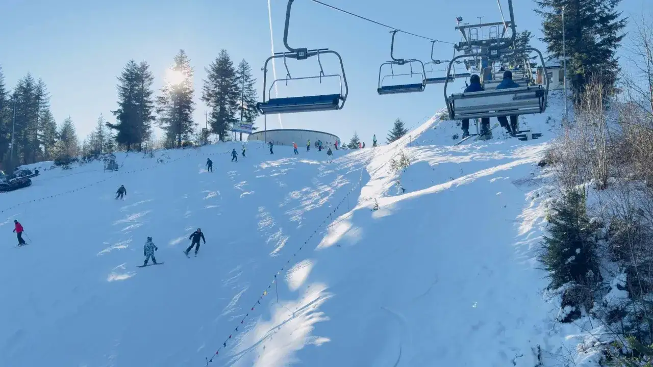 Słoneczny dzień na stoku w Myślenicach. Narciarze i snowboardziści szusują po ośnieżonych górach, a kolejka linowa wwozi kolejnych na szczyt.