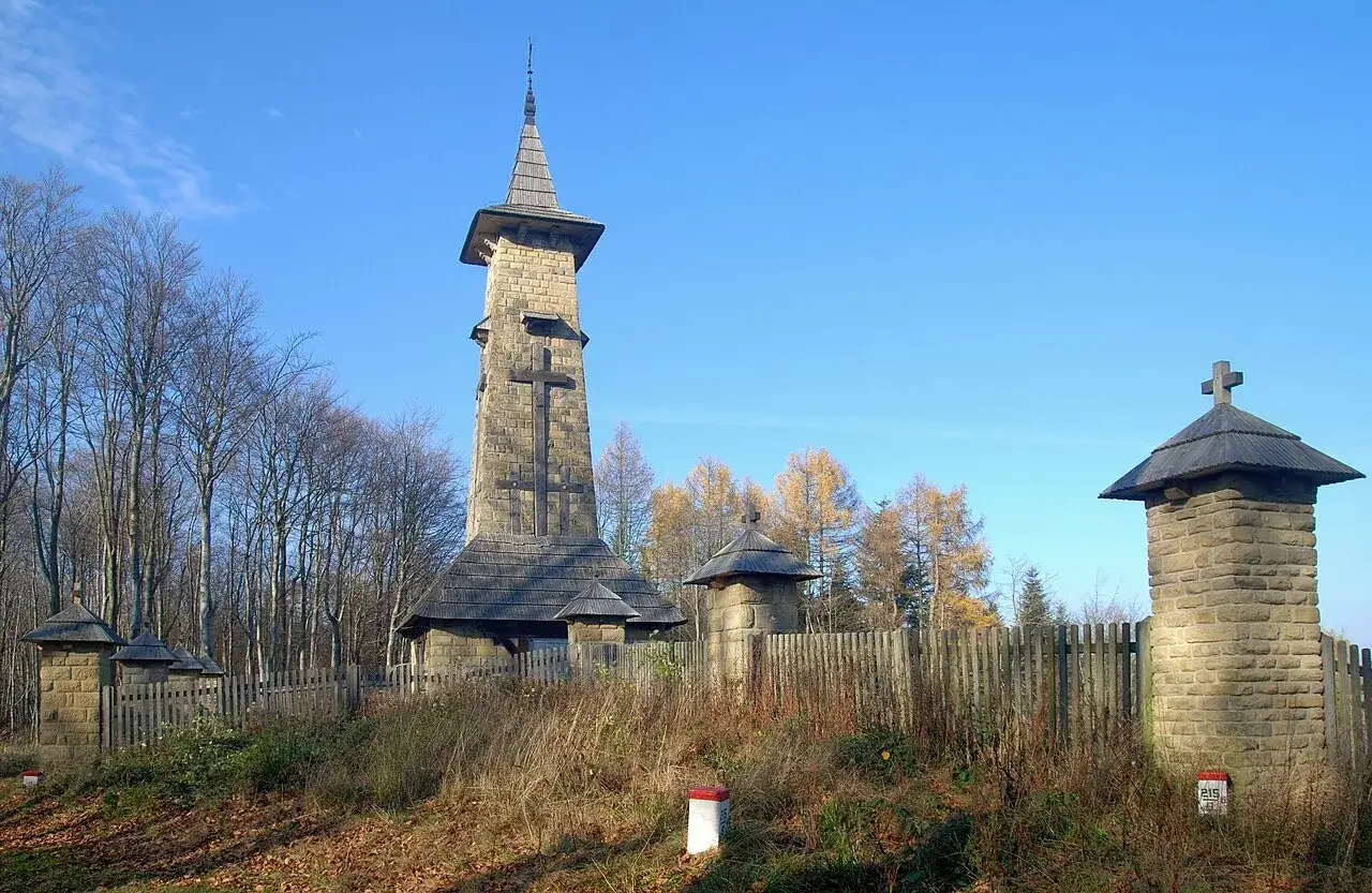 Kamienna wieża z krzyżem i drewniane kapliczki, otoczone płotem i drzewami. To jedne z regietów atrakcje w jesiennym krajobrazie.