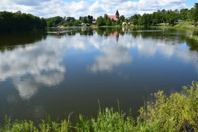 Spokojne jezioro na Kaszubach, odbijające chmury. W oddali widać łódkę i kościół. Idealne miejsce na wakacje.
